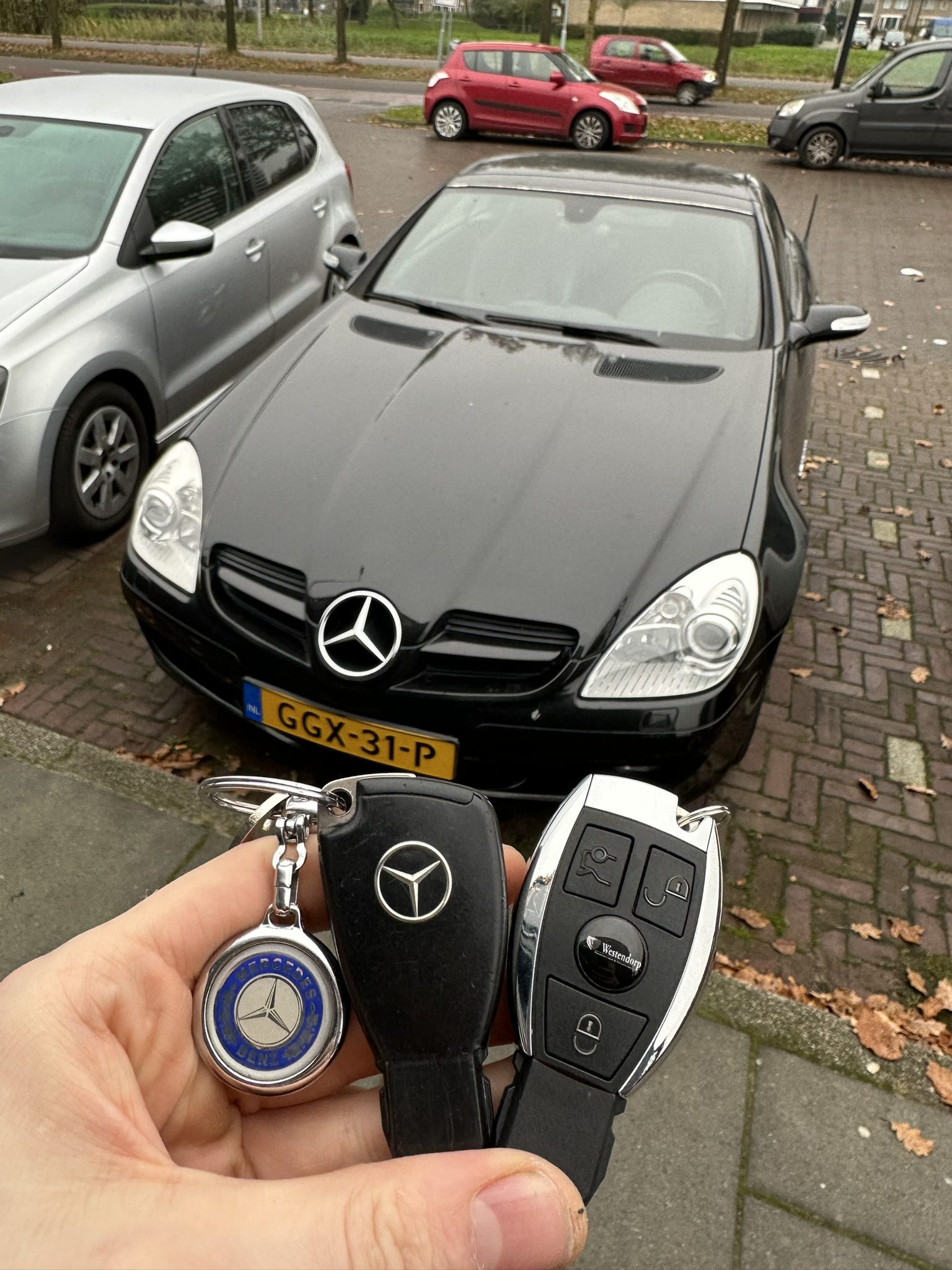 Mercedes-sleutel met sleutelhanger in de hand, met de zwarte Mercedes SLK op de achtergrond