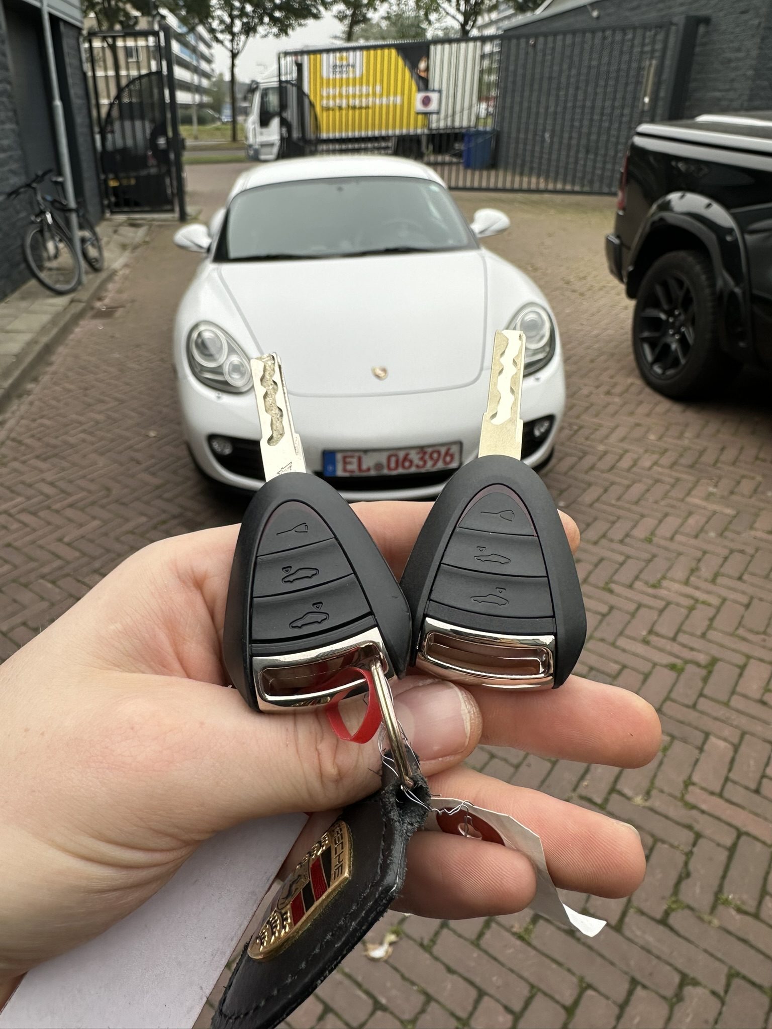Twee Porsche-sleutels in de hand, met de witte Porsche Boxster op de achtergrond