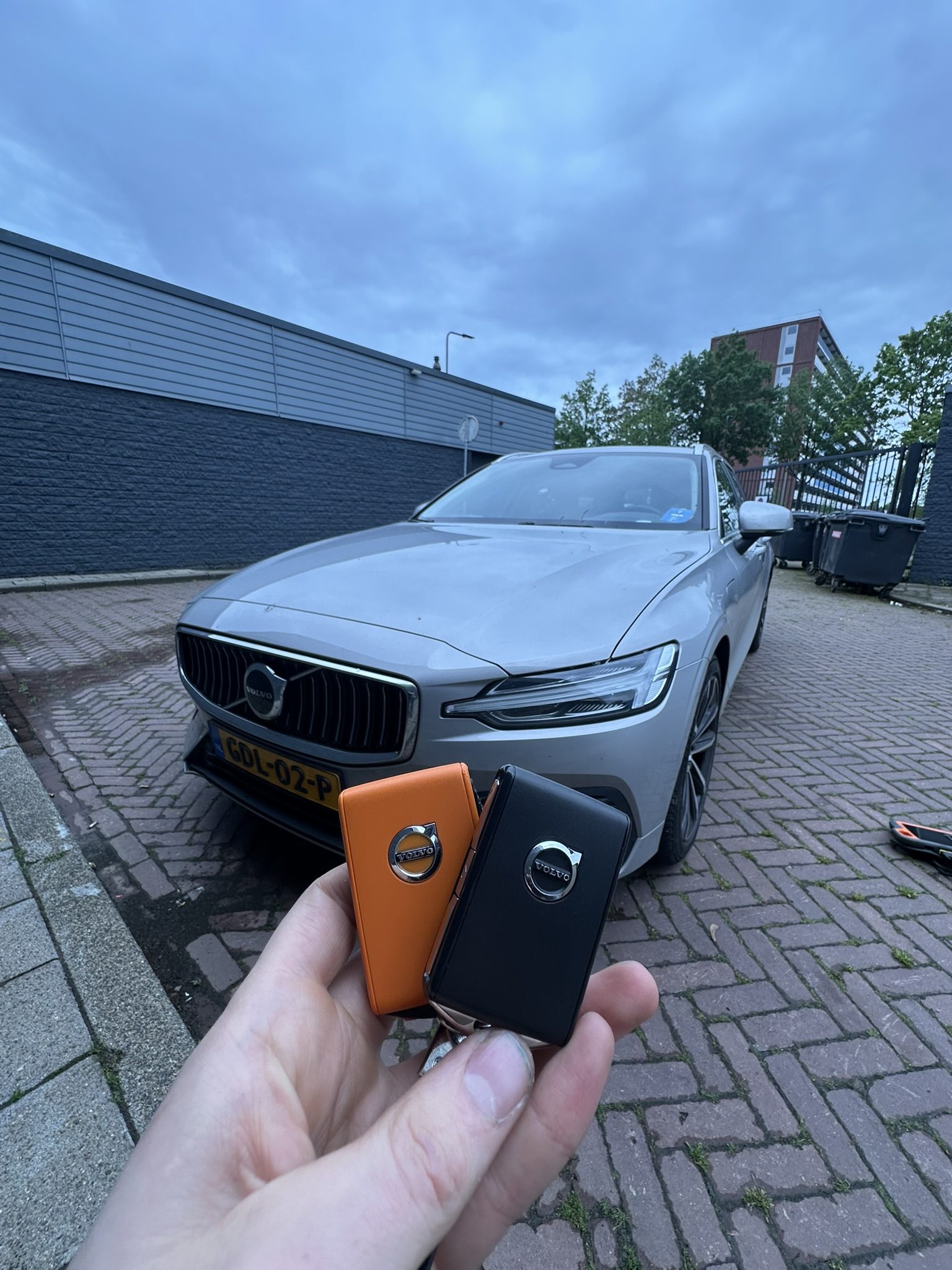 Volvo-sleutel met oranje hoes in de hand, met de grijze Volvo V60 op de achtergrond